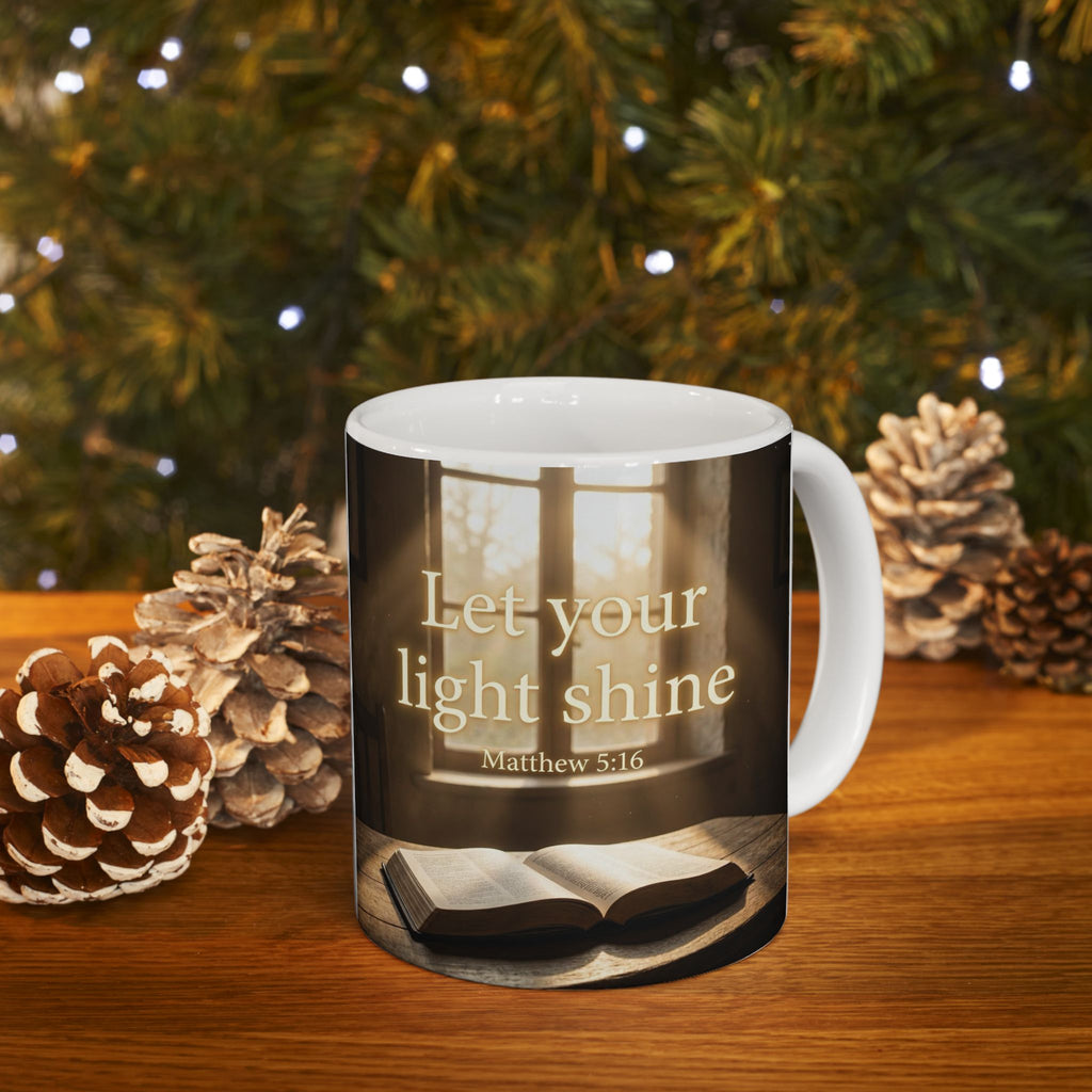 A white ceramic mug with a printed graphic of a Bible on a table in front of a window, featuring the text 'Let your light shine Matthew 5:16'. The mug is placed on a wooden surface with pinecones and a blurred Christmas tree background.